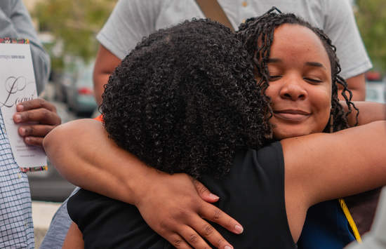 Two female students embracing each other while a background figure holds a LOQUI pamphlet