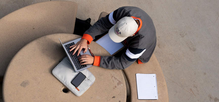 Student typing on his laptop, empty notebook next to him