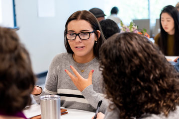 Seaver student in a gray sweater, talking with her hands to women seated at her table