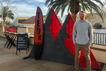 David Bird standing alongside a red and black surfboard and mountain peaks-artwork he created for TEDx