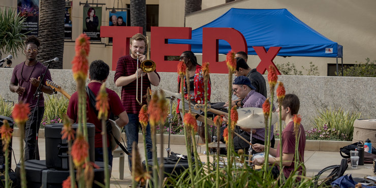 Red TEDX logo standing upright with student musicians playing the drums, saxophone, and guitar in front of it