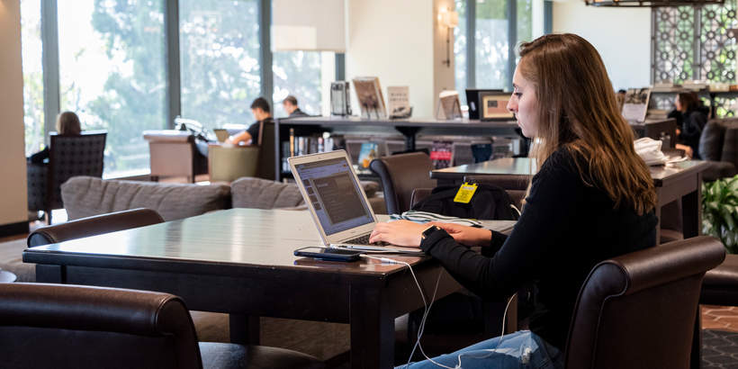 Female student sitting at a high desk in Payson library typing on her laptop