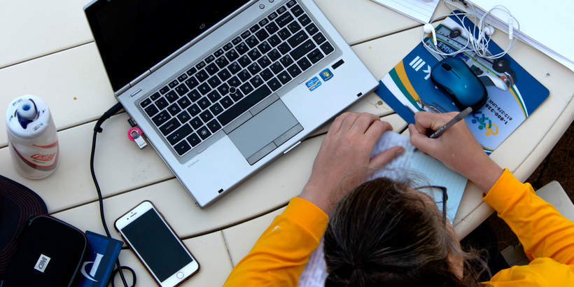 Student in a yellow sweater writing in a notebook on a table with a laptop and cellphone beside her