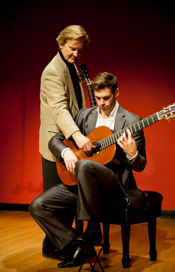 Chris Parkening adjusting his student's guitar strings while on stage. Student is seated, cradling guitar.