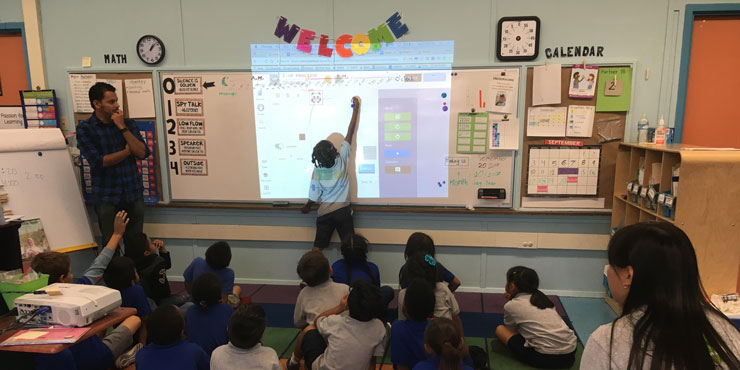 Student teacher looking at whiteboard as a student writes on it, his peers are seated on the carpet behind him