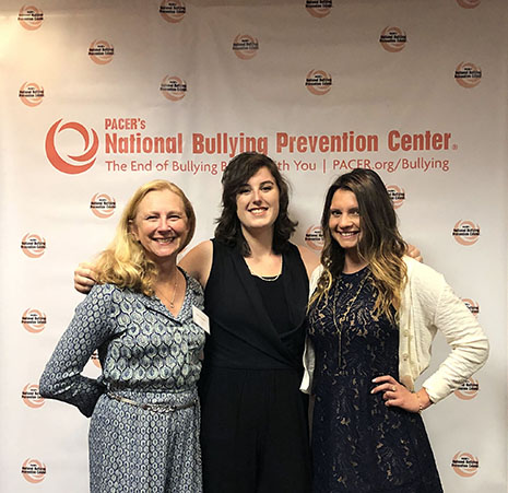 Pepperdine students Tiffany Hall and Anne Mummery stand with Judy French in front of backdrop with PACER's National Bullying Prevention Center's logo after accepting award.